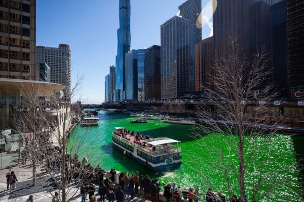 A photograph captures a festive scene in downtown Chicago during St. Patrick’s Day celebrations, where the Chicago River is dyed a vibrant green. Boats cruise along the river, carrying passengers enjoying the event. A crowd gathers along the riverwalk, many dressed in warm clothing, watching and taking photos. The city’s skyline, featuring modern skyscrapers and iconic architecture, rises in the background under a clear blue sky. Leafless trees in the foreground frame the lively scene.