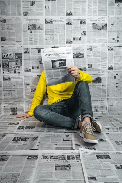 Person in a bright yellow hoodie and jeans sits on a floor covered with newspaper pages, holding an open newspaper in front of their face so it hides their identity, with a wall of newspapers behind them. Photo by Egor vikhrev IFdQ6ea7r0s unsplash.