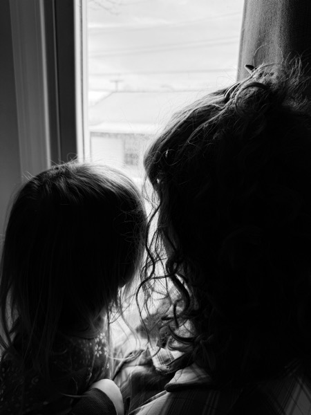Black‑and‑white photo of an adult and a small child with long hair seen from behind, standing close together at a window and looking out at a snowy yard and building.
