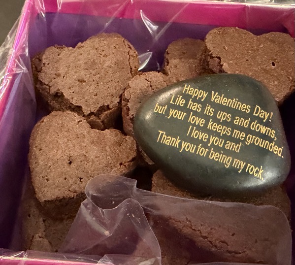 A close-up of a Valentine’s Day gift box filled with heart-shaped chocolate brownies and a smooth black stone engraved in gold text that reads, “Happy Valentines Day! Life has its ups and downs, but your love keeps me grounded. I love you and thank you for being my rock.”