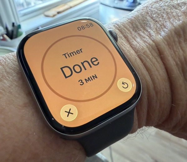IMG_2362.jpeg Close-up of a person’s wrist wearing an Apple Watch displaying a bright orange screen that reads “Timer Done 3 MIN,” with cancel and repeat icons, in a cluttered home office setting.