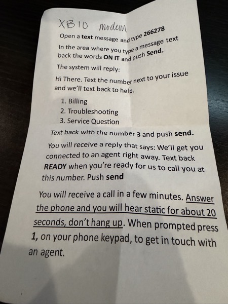 Photo of a printed instruction sheet on a dark table with “XB10 modem” handwritten at the top, explaining how to text 266278 for billing, troubleshooting, or service questions, and detailing that after replying “READY,” the user will receive a call, hear about 20 seconds of static, and then must press 1 to reach an agent.