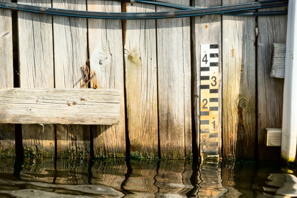 Water level gauge mounted on a weathered wooden dock wall, with black and yellow measurement markings partly submerged in calm, reflective water.