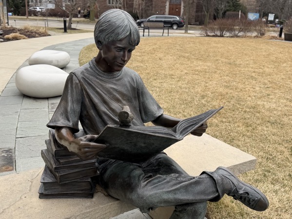 Bronze statue of a child sitting on an outdoor stone bench with legs crossed, reading an open book that rests on their lap, a small bird perched on the top edge of the book, and a stack of books beside them, with a paved walkway, grass lawn, and buildings in the background.