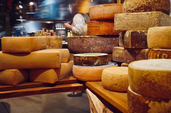 A rustic indoor display features a wide variety of artisanal cheese wheels and blocks stacked tightly on wooden shelving. On the left, smooth, rectangular orange-brown blocks are piled horizontally. On the right and center, large round wheels of varying sizes are stacked vertically, displaying diverse rinds—ranging from textured dark brown and dusty gray to smooth ochre and patterned beige. Photo by Azzedine rouichi YW_5rJvAdKw unsplash.