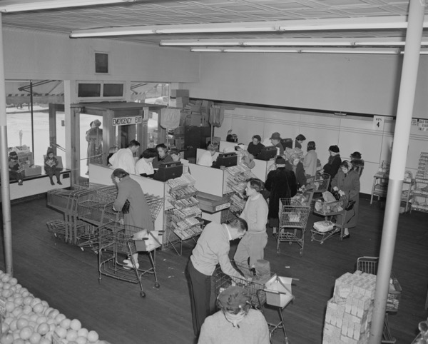 An high angle, wide shot of an old black and white photograph captures the interior of a grocery store where shoppers wait in line at checkout stands.