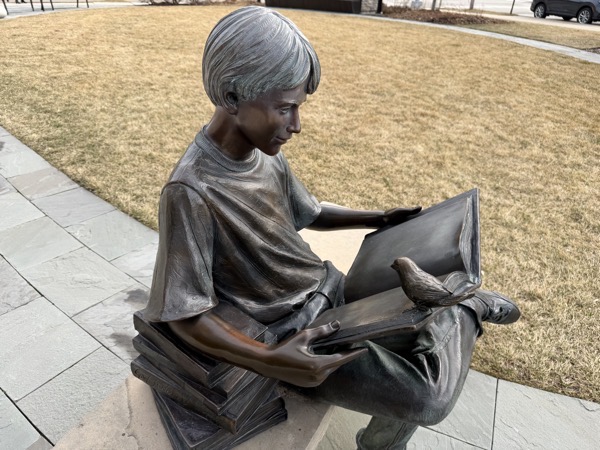 A close-up photograph captures a bronze statue of a young boy sitting on a stone bench outdoors, absorbed in reading a book.