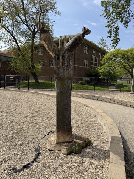 A large wooden sculpture carved from a tree trunk stands in the center of a gravel-covered dog park. The sculpture is shaped like a giant fork skewering a sausage. The base of the sculpture is a wide, flat tree stump. Behind the sculpture is a two-story brick building with several windows, and to the sides are lush green trees under a clear blue sky. A low stone wall and a paved path curve around the gravel area. A black chain-link fence separates the park from the building. Two tennis balls lie on the gravel to the left of the sculpture.