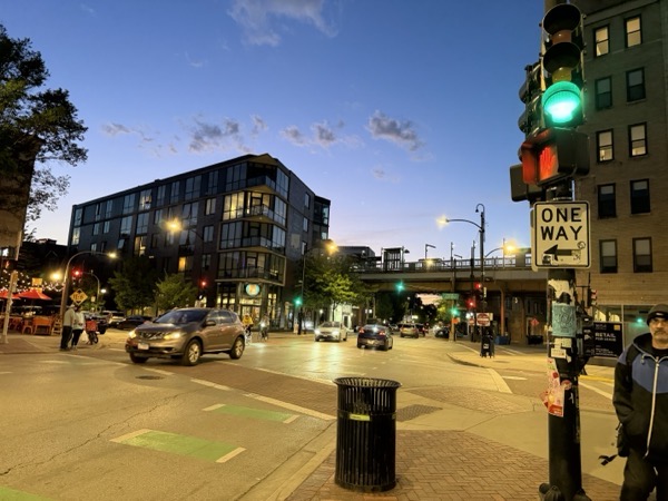 A wide-angle, low-light shot captures a busy urban intersection at twilight in Chicago. In the foreground, a prominent black traffic light pole features a glowing green light and a "One Way" sign pointing left, heavily adorned with stickers. A black metal trash can sits on the brick sidewalk nearby.