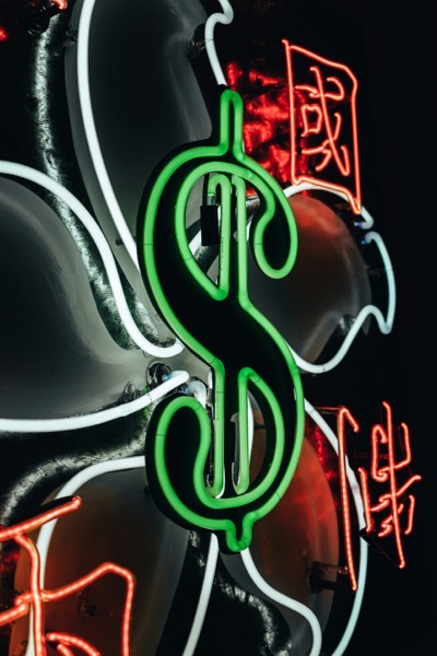 A close-up photograph of overlapping neon signs illuminating a dark background. In the foreground, a prominent bright green neon sign is shaped like a dollar symbol ($\$$) by Aedrian salazar Wy7Gy8ZcW1M unsplash.