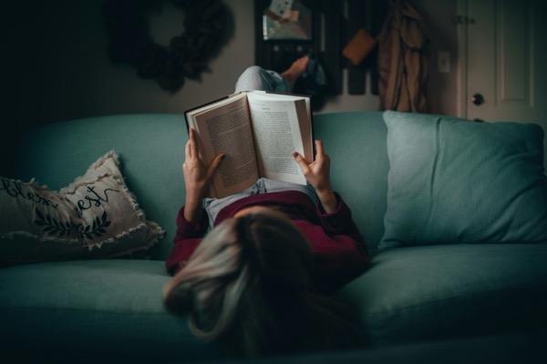 An overhead, first-person perspective shows a person lying on their back on a teal couch, holding open a large book to read. The person's head is closest to the foreground, with long, blonde-streaked hair spilling downward. They are wearing a deep maroon, long-sleeved sweater and light grey sweatpants. Image from Matias north v8DSLoY80Xk unsplash.