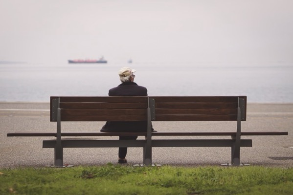 an elderly person wearing a dark jacket and a light-colored baseball cap sits alone on a long wooden park bench with metal support brackets. The bench faces a vast, calm body of water under a hazy, overcast sky. In the far distance, a large cargo ship is faintly visible on the horizon. Low green grass lines the foreground.