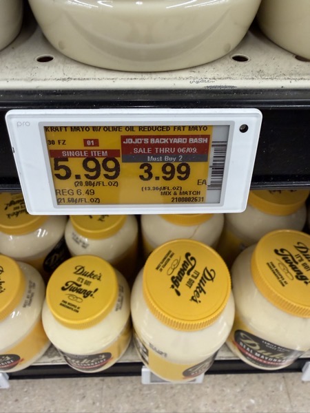 A grocery store shelf stocked with jars of Duke's Mayonnaise under a digital price tag. The yellow tag displays product details for "Kraft Mayo w/ Olive Oil Reduced Fat Mayo 30 fl oz," showing a single item price of $5.99 (regularly $6.49) and a "Jojo's Backyard Bash" sale price of $3.99 each when buying two (sale thru 06/09). The shelf below contains multiple jars of Duke's Real Mayonnaise with distinctive yellow lids reading "Duke's - It's Got Twang!"