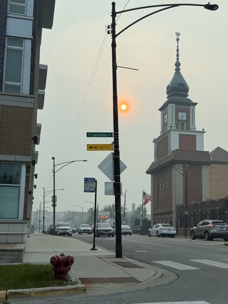A hazy street scene in Chicago looking down a road with cars. A tall street light pole with a green "N Laramie Av" sign stands in the foreground, with the prominent clock tower of the Copernicus Center visible on the right against a dim, glowing sun obscured by thick smoke from Canadian wildfires