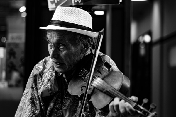 Guillaume gryn_dvs kv5v3 Yh _M unsplash. Black-and-white close-up of an older man playing a violin, wearing a light fedora with a dark band and a patterned shirt, focused with an expressive, open-mouthed expression as he draws the bow across the strings.
