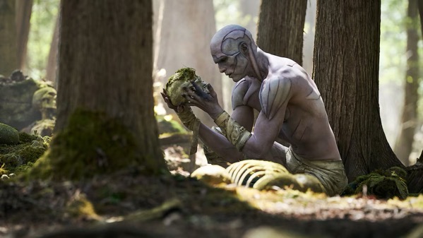 A publicity still from Guillermo del Toro's Frankenstein featuring a sunlit forest surrounded by tall tree trunks, a pale, muscular humanoid creature with light purple skin and ridge-like markings on its head and shoulders crouches on the ground. The figure wears cloth wraps around its forearms and waist, and holds a mossy human skull close to its face with a somber expression. Partial human skeletal remains, including a rib cage, rest on the mossy forest floor in front of it.