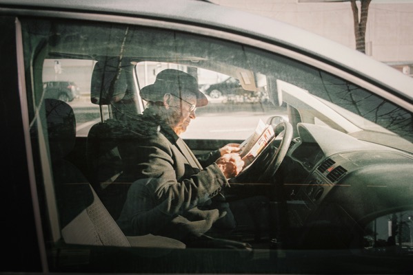 samuel-couto-bKBOP9pn1K0-unsplash.jpg A side-profile view shot through a car window showing an elderly man wearing a fedora hat, glasses, and a dark jacket sitting in the driver's seat. He is holding and reading a newspaper open near the steering wheel. Reflection from the street is subtly visible on the glass, giving the image a muted, film-like aesthetic. Image by Samuel Couto bKBOP9pn1K0 unsplash.