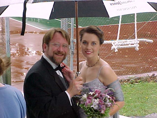 umbrella.jpg a couple on their wedding day standing under an umbrella in front of a wet tennis court