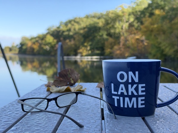 a blue mug with the words "ON LAKE TIME" printed in white, sitting on a wet wooden dock alongside a pair of black-framed glasses and a few fallen autumn leaves. In the background, a calm lake reflects the surrounding trees under a clear blue sky.