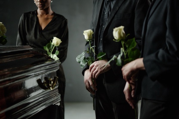 shutterstock_2684119373.jpg Black woman and two Caucasian adults standing beside coffin holding roses, participating in funeral ceremony, somber expressions visible, partial faces and torsos shown Shutterstock 2684119373.